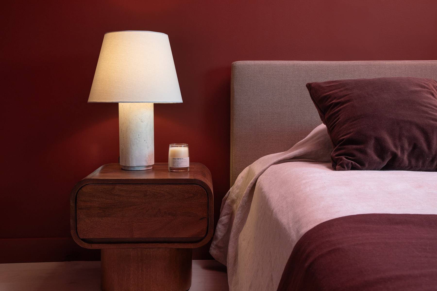 One bedroom detail showing corner of bed and nightstand with lamp and candle, maroon tones