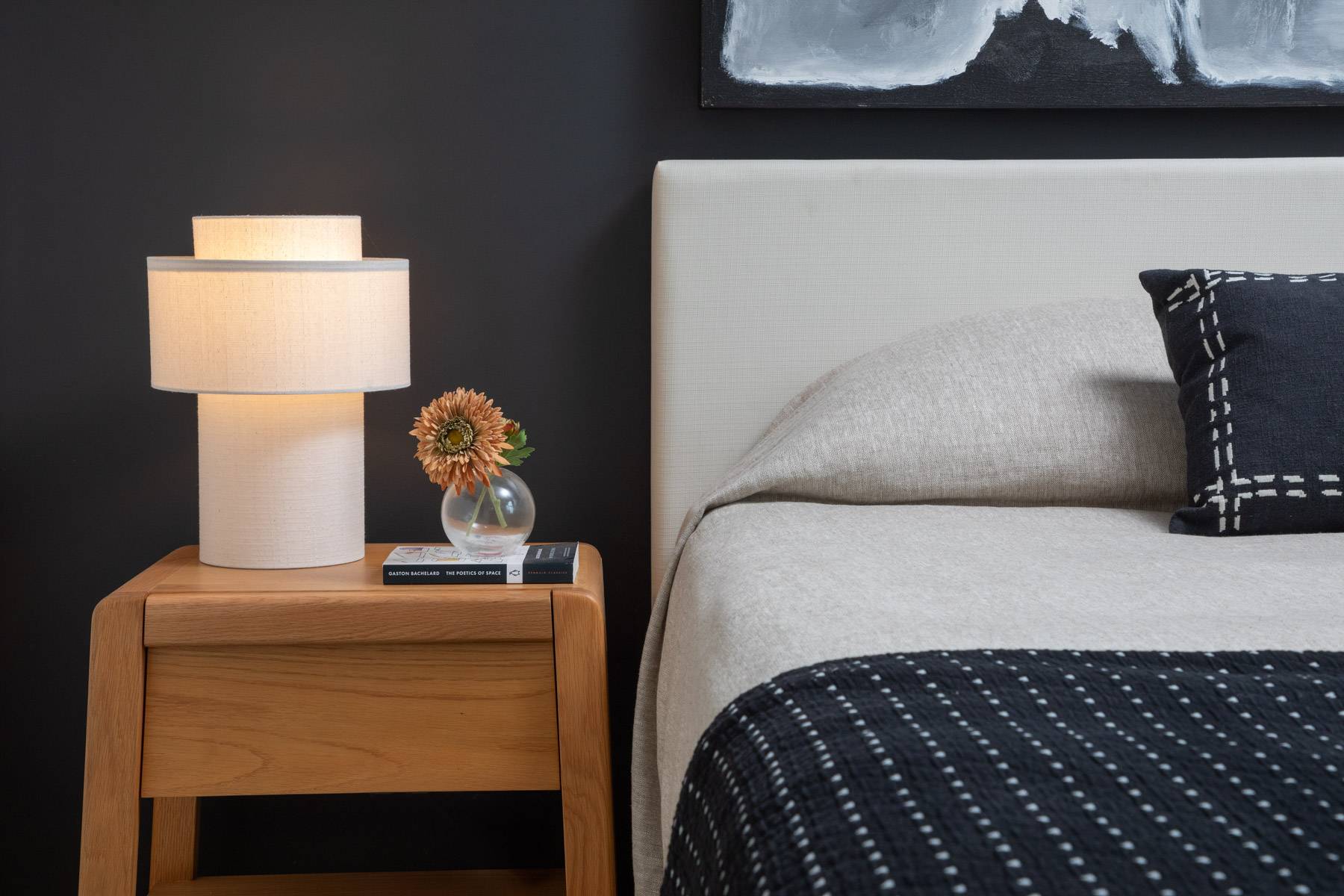 Studio bedroom detail showing bed and nightstand with lamp and flower in glass vase on book