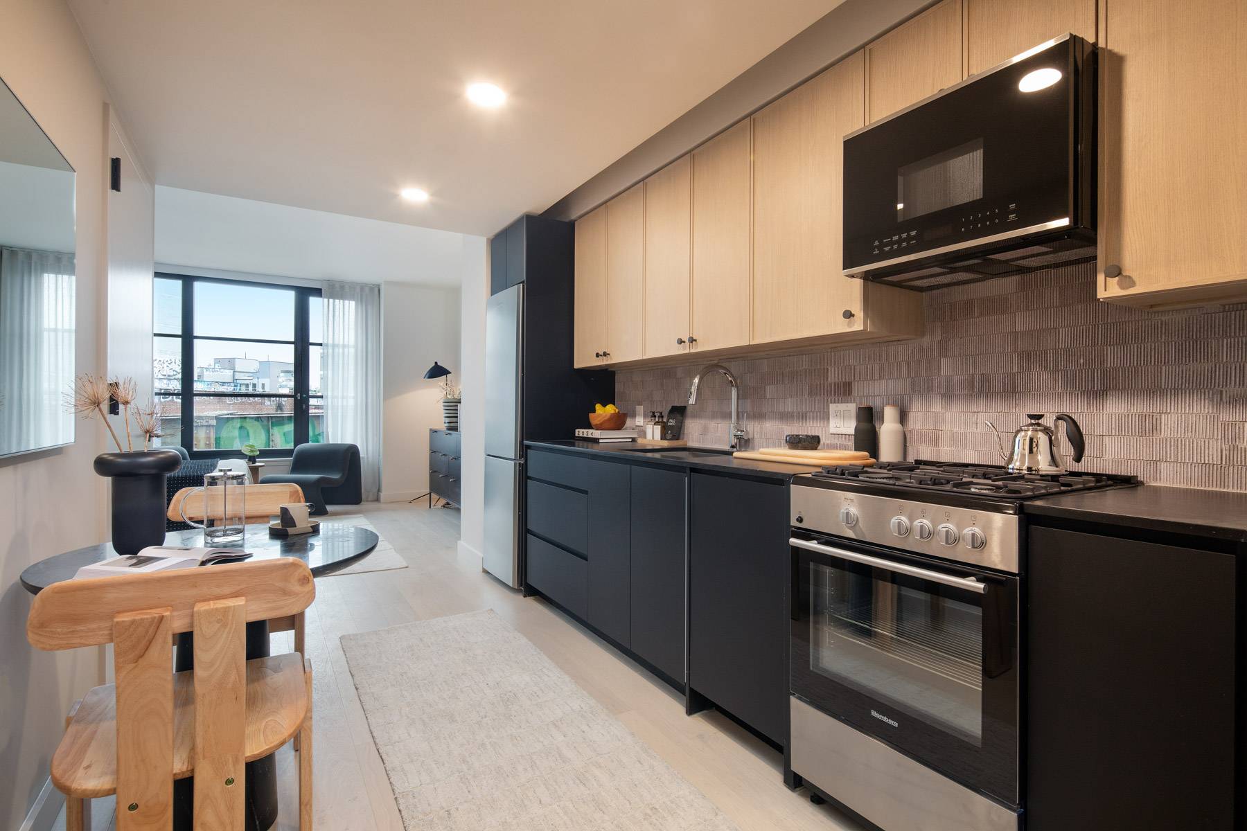 Studio galley kitchen with black and stainless appliances, light upper cabinets, dark lower cabinets, light flooring and eat in table for two